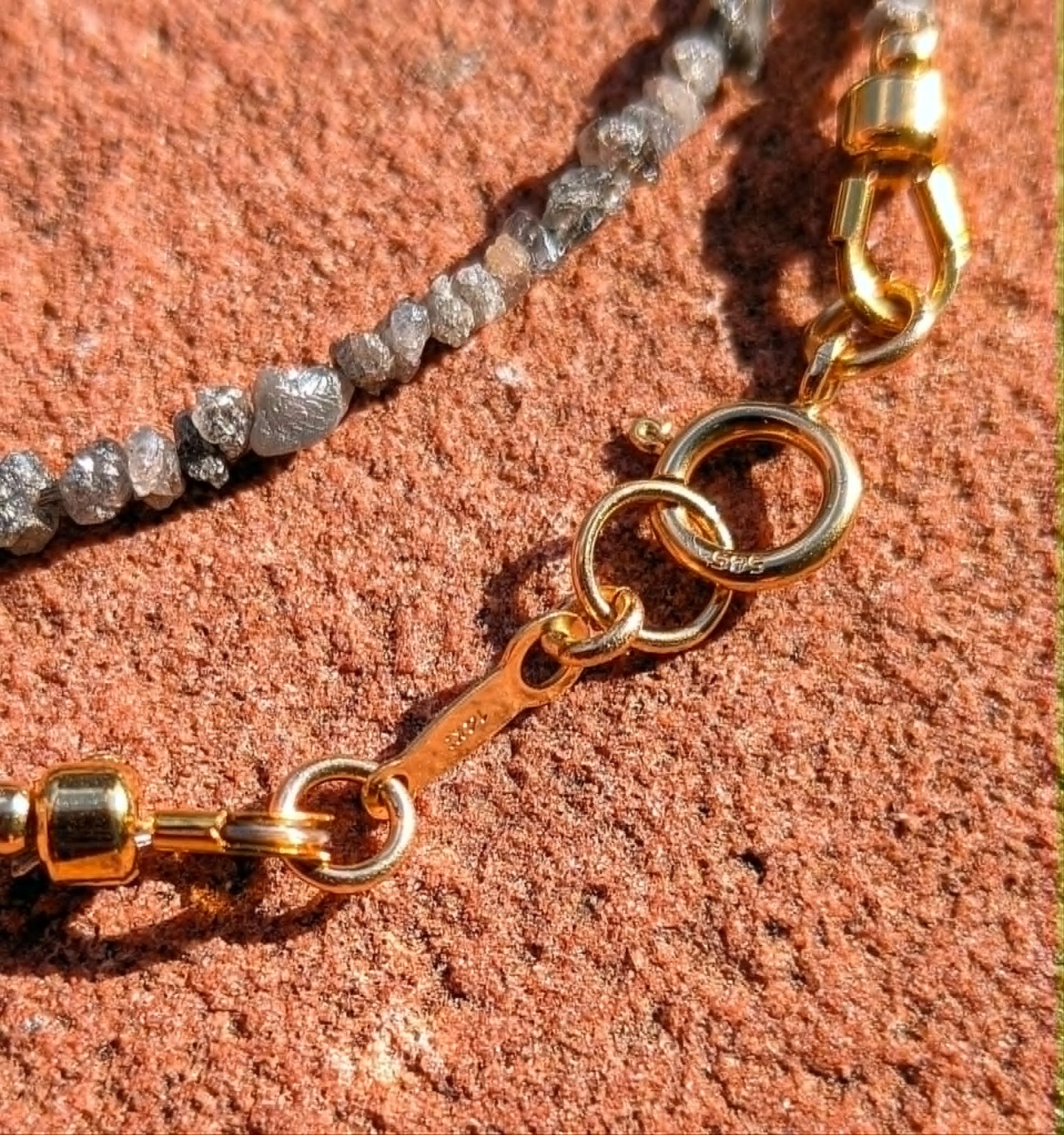 Close-up of a beaded chain necklace with clear faceted diamond beads and gold metal rings and clasp, laid on a textured reddish-brown stone surface.