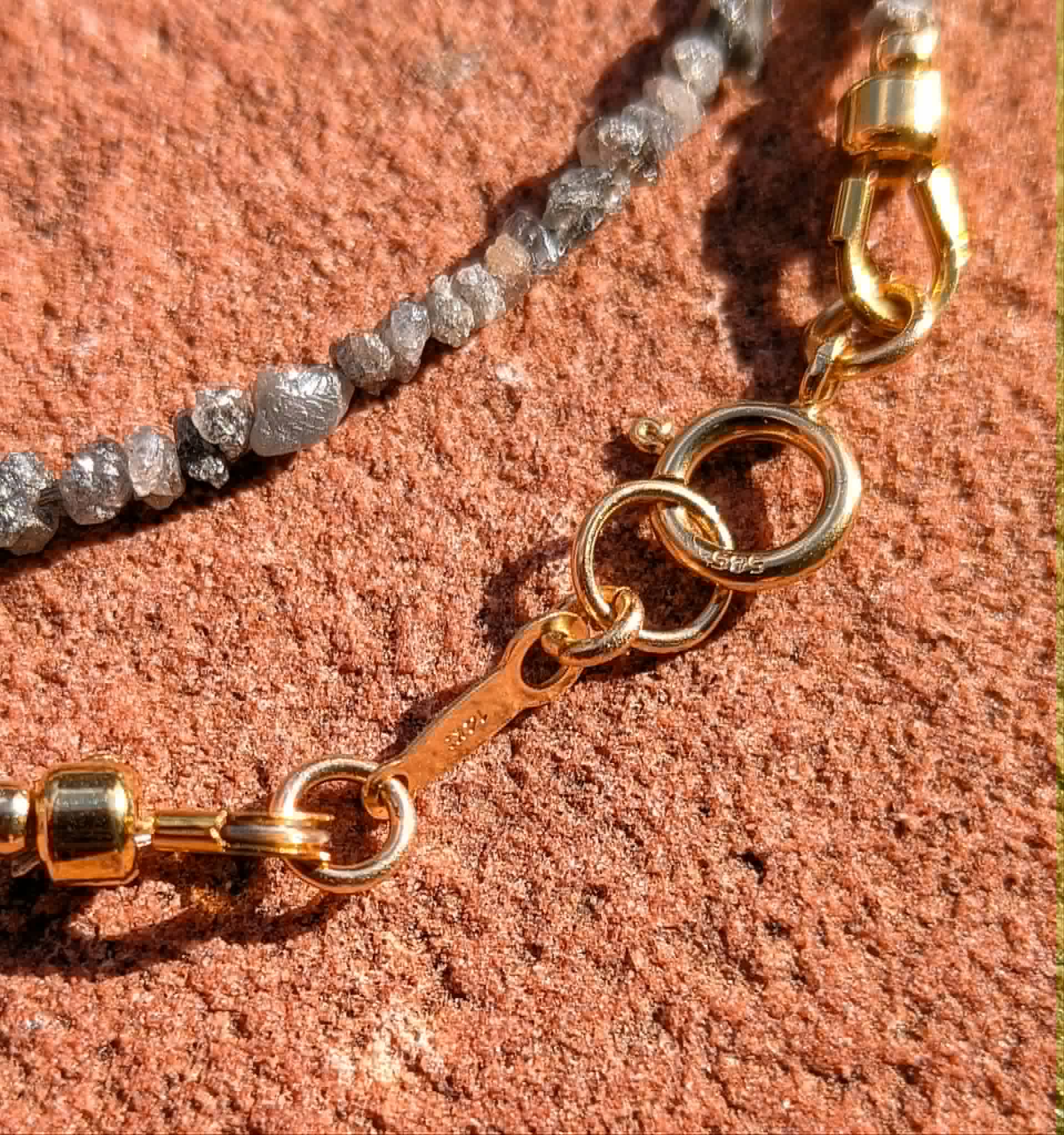 Close-up of a beaded chain necklace with clear faceted diamond beads and gold metal rings and clasp, laid on a textured reddish-brown stone surface.