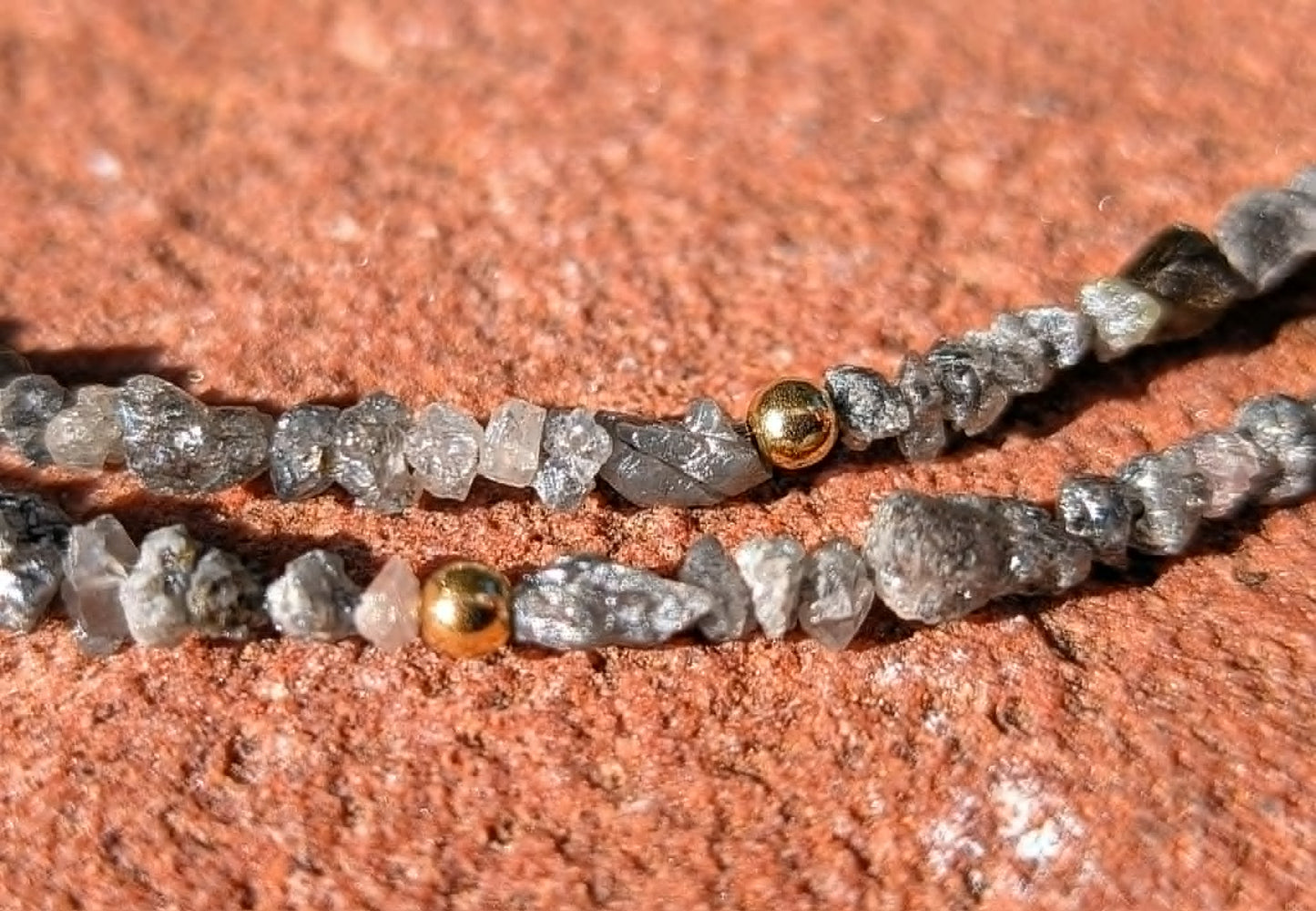 A close-up of a beaded strand featuring rough, translucent gray uncut rough diamond beads and gold spacer beads, laid on a textured reddish-brown stone surface.