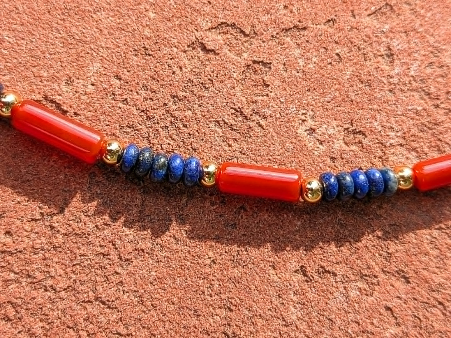 A close-up of a beaded necklace featuring vibrant red cylindrical carnelian beads, deep blue lapis lazuli rondelle beads, and small gold beads, laid on a textured reddish-brown surface.
