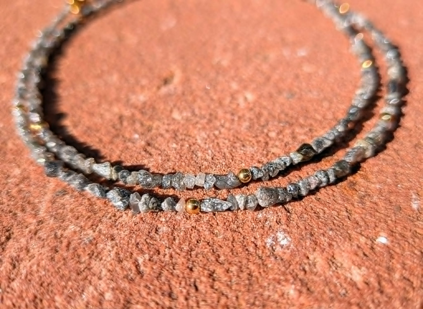 A doubled strand of a beaded necklace made of small gray and black rough uncut diamond beads with a few metallic gold accent beads, displayed on a textured reddish-brown surface.