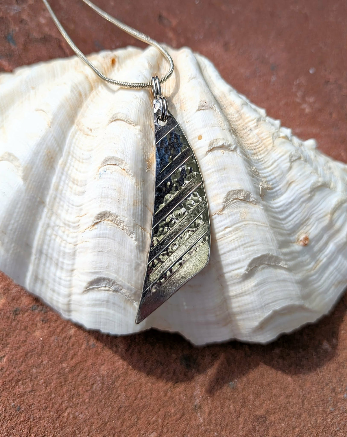 A silver sail-shaped pendant necklace with a textured surface, resting on a large white seashell against a reddish-brown sandy background.