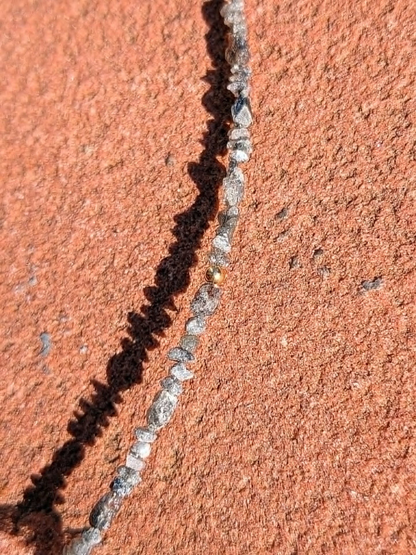 A close-up of a textured terracotta or rust-colored surface with a narrow dark and light gray strand of rough uncut grey diamond beads running diagonally through the frame.