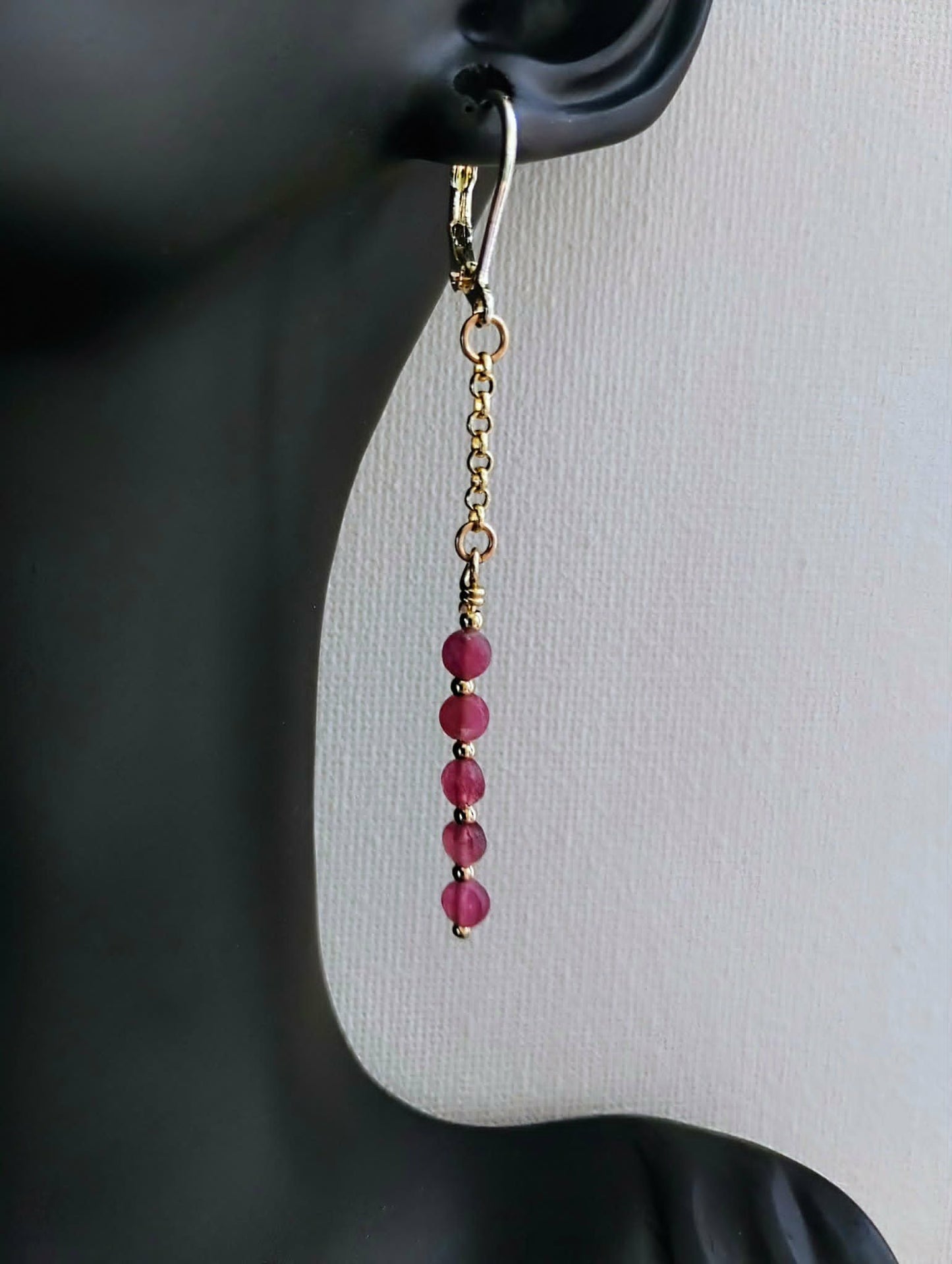 A single dangling earring displayed on a black mannequin ear against a light gray background. The earring has a gold-toned fishhook and chain with a vertical drop of faceted pink beads.