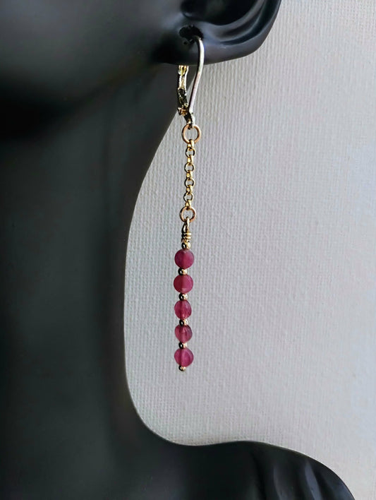 A single dangling earring displayed on a black mannequin ear against a light gray background. The earring has a gold-toned fishhook and chain with a vertical drop of faceted pink beads.
