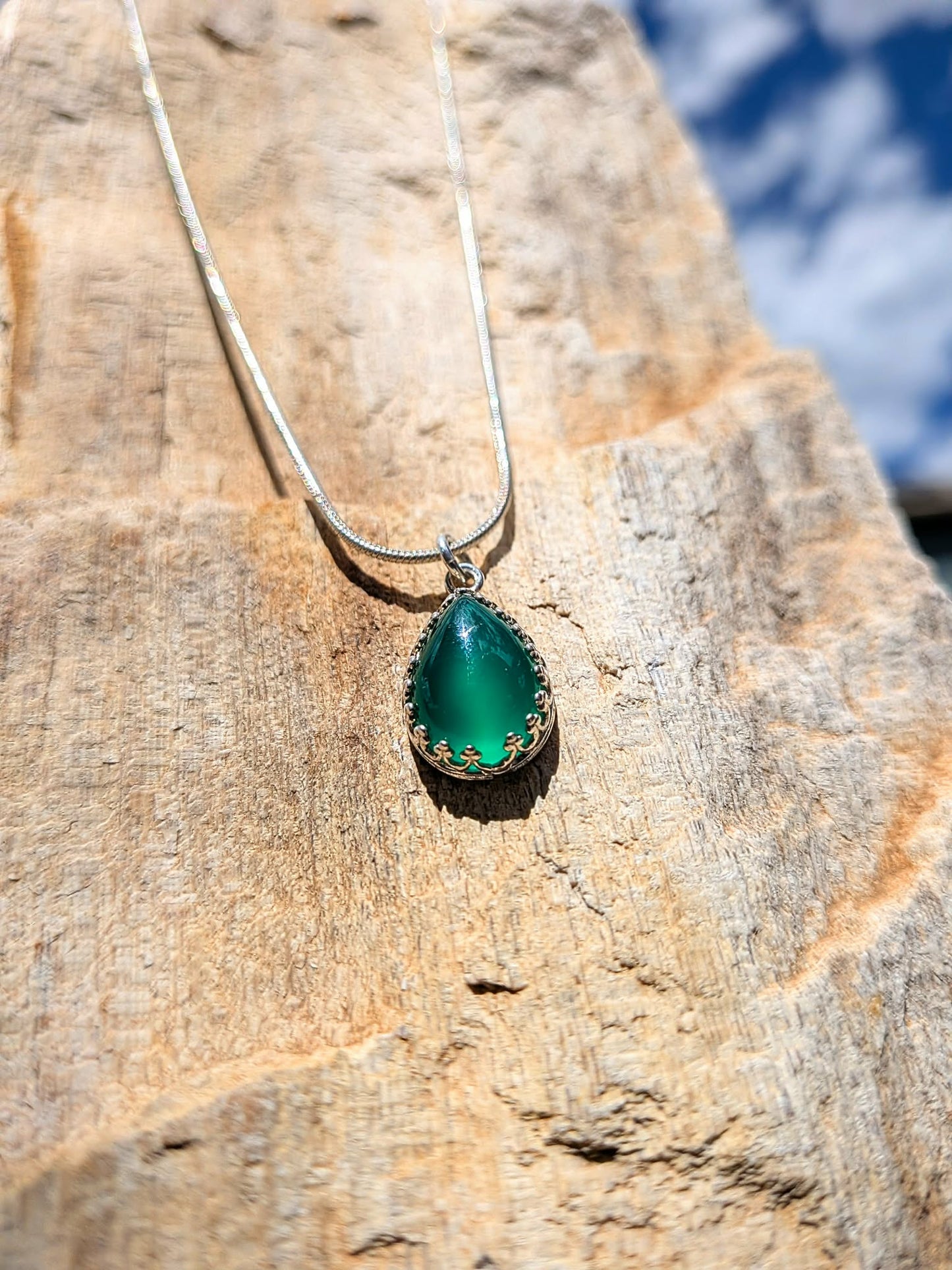 A silver necklace with a teardrop-shaped green gemstone pendant, resting on a textured tan rock outdoors.