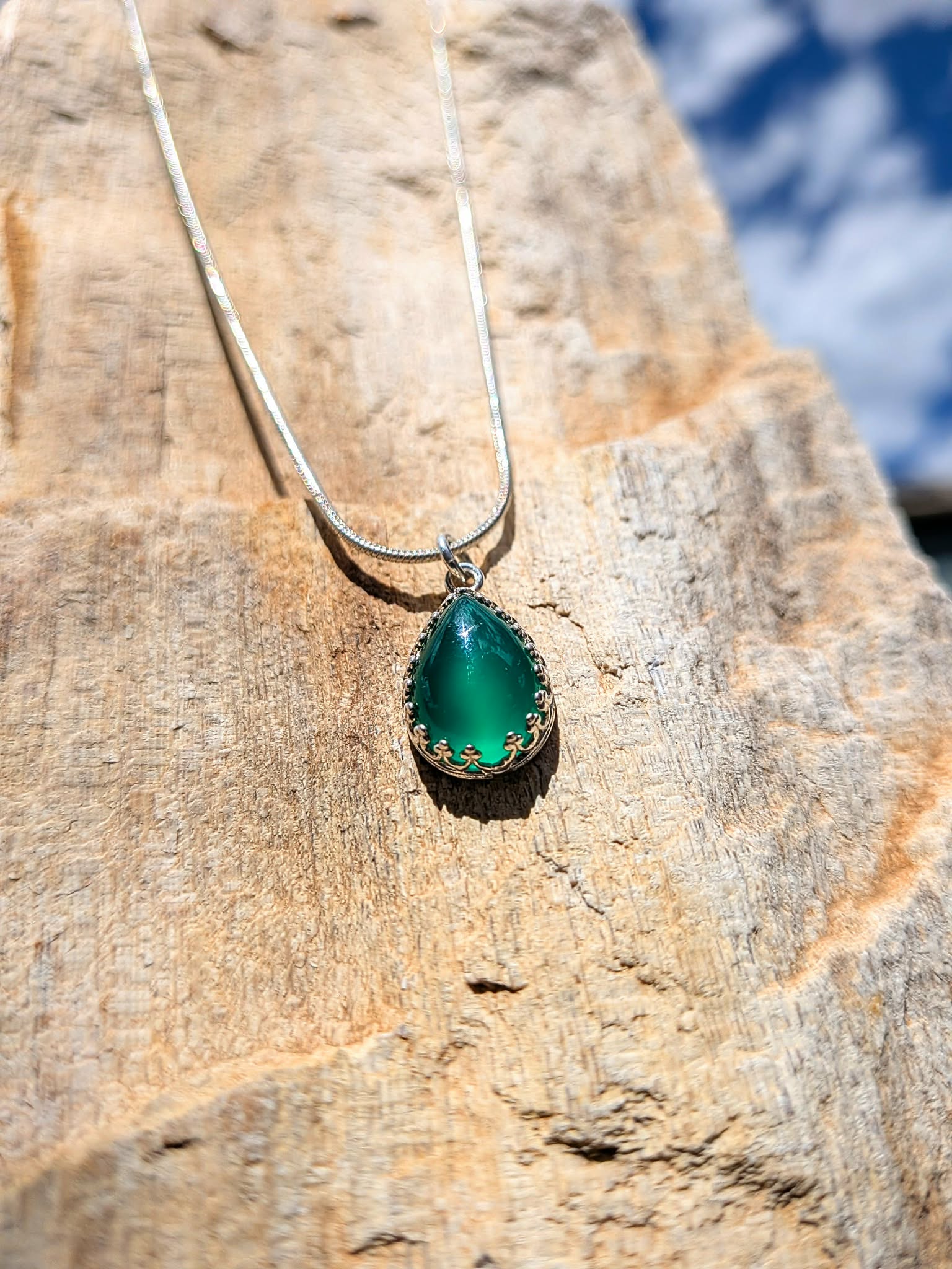 A silver necklace with a teardrop-shaped green gemstone pendant, resting on a textured tan rock outdoors.