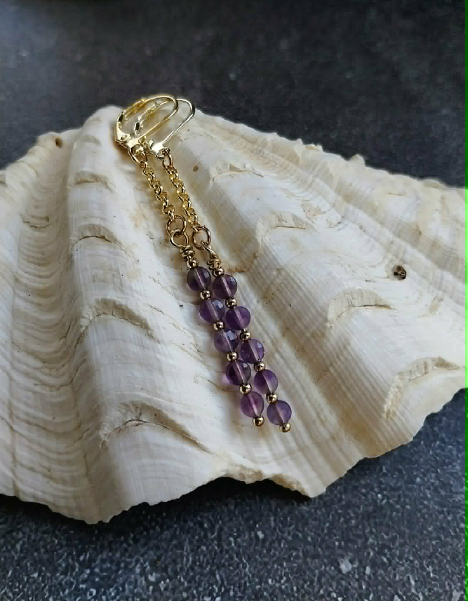 A gold-toned metal keychain or bag charm with a series of purple faceted beads, displayed on a large white seashell against a dark textured background.